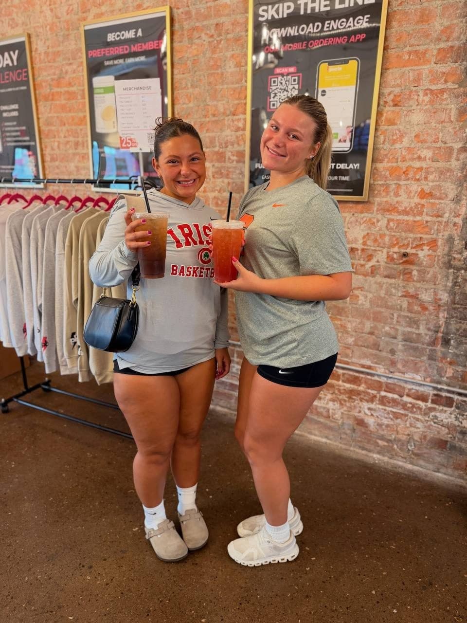 Two athletes smiling with their loaded teas in front of the club's brick wall and merch rack