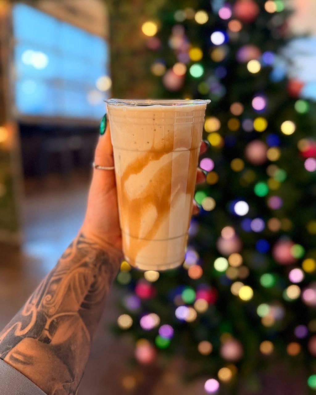 A caramel-swirled protein shake held up in front of a lit Christmas tree
